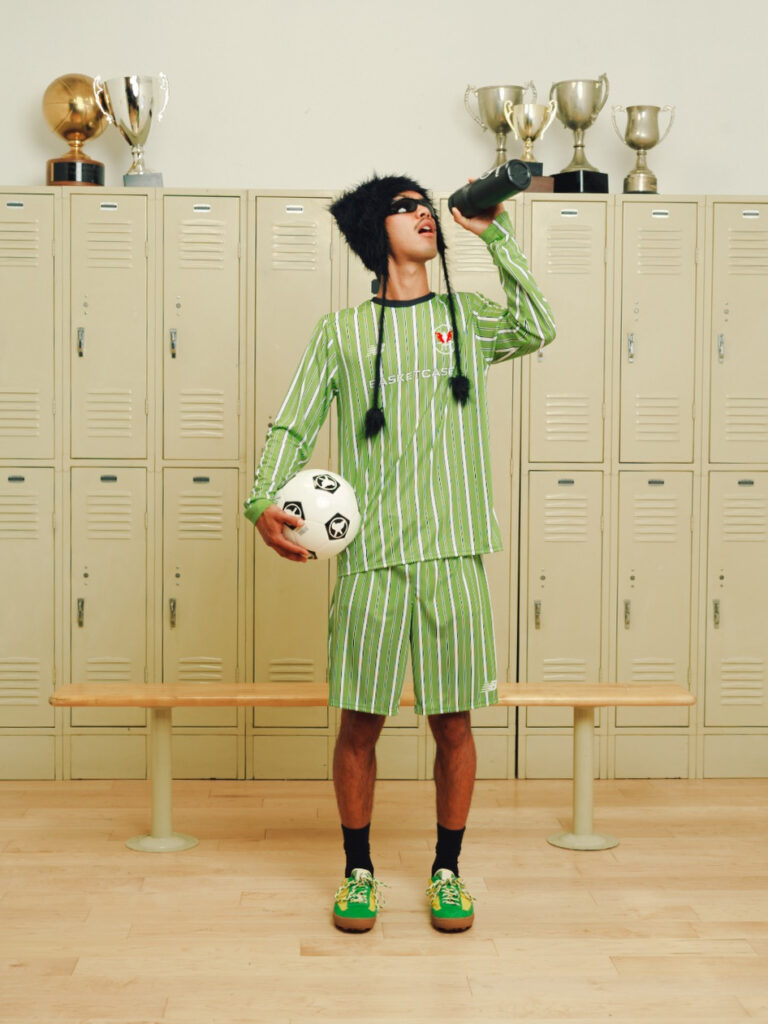 Person in a green striped soccer uniform drinks from a bottle while holding a soccer ball in a locker room with trophies on top of the lockers.