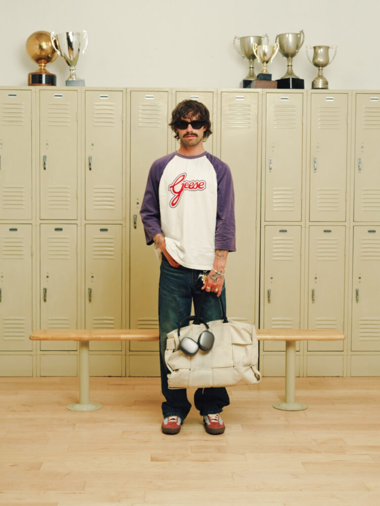 Man with sunglasses stands in a locker room facing the camera, holding a beige duffel bag with headphones and wearing a white and purple Geese shirt.