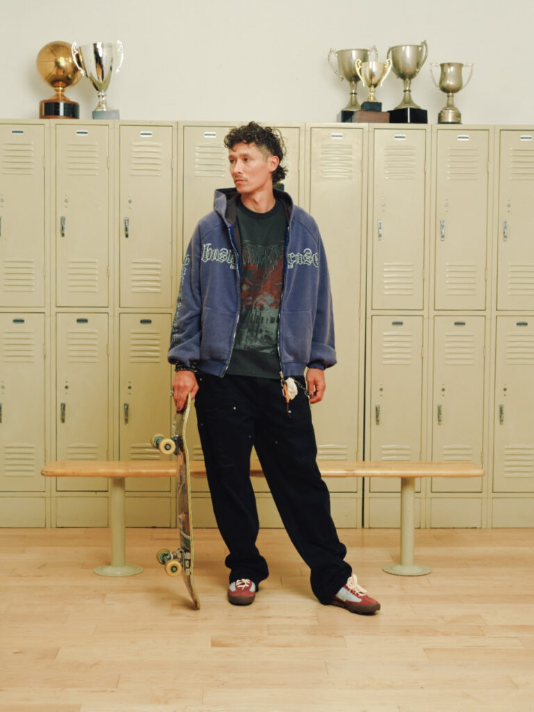 Young man in a blue hoodie and dark pants stands with a skateboard in front of beige lockers, trophies on top shelves in a school locker room.