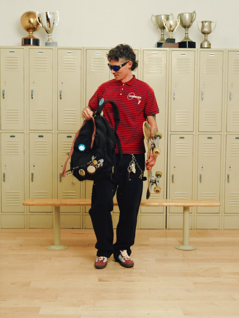 Skateboarder in a red striped polo and sunglasses stands by beige lockers, carrying a skateboard and backpack.