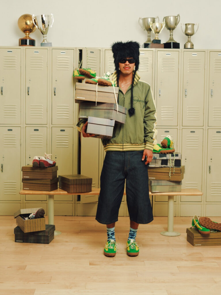 Person in a black furry hat and sunglasses stands in a locker room, balancing a stack of shoeboxes and sneakers.