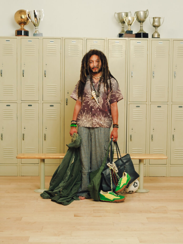 Person with dreadlocks wearing a tie-dye shirt and checkered pants stands in a school locker room, holding a green jacket and a black bag with a soccer ball nearby on the floor/bench behind them.