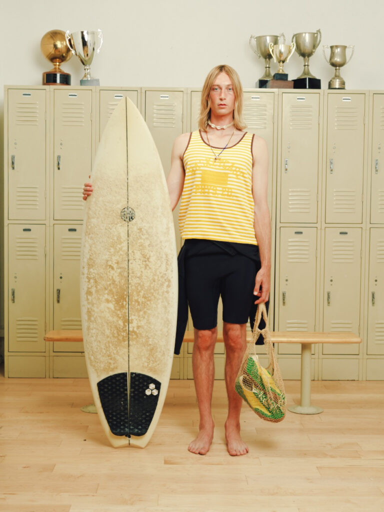 Young man with blond hair in a yellow striped tank top and black shorts stands barefoot in a locker room, holding a surfboard and a mesh bag.