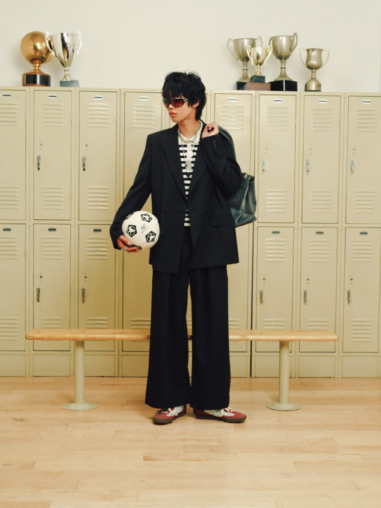Person in a black suit and striped shirt holding a soccer ball, standing in front of beige lockers with trophies on top shelf.