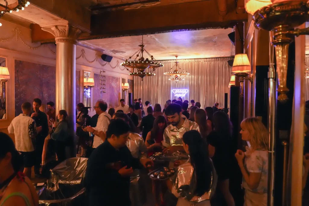 Crowded bar/club interior with warm lighting, ornate columns, chandeliers, and people mingling and chatting at tables and the bar area.