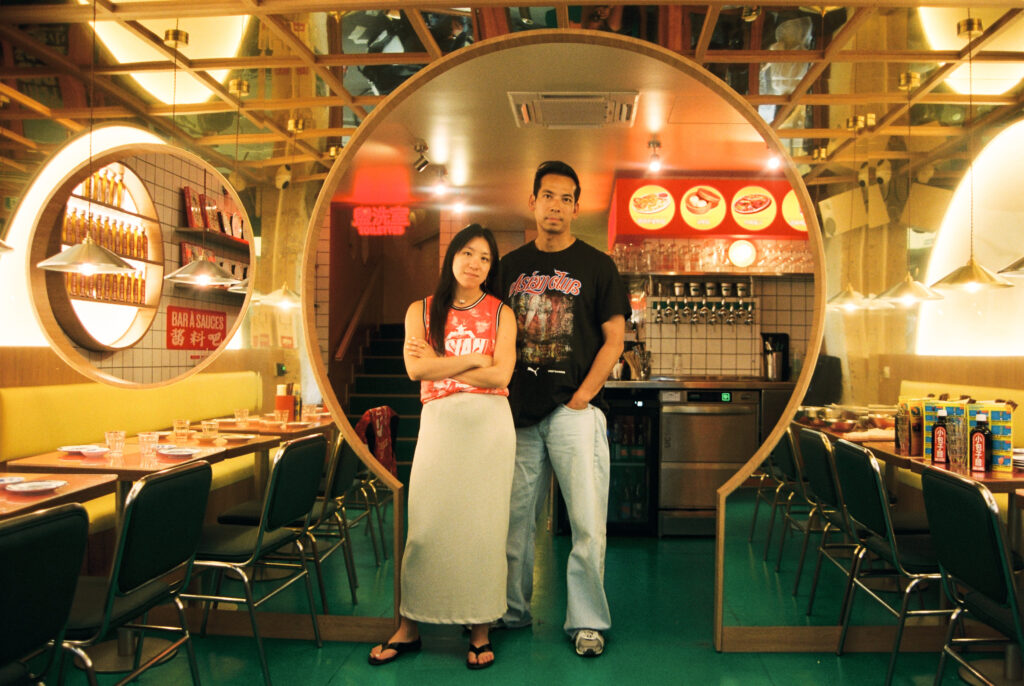 Two people stand together in a retro-style diner, framed by large circular mirrors and warm yellow lighting behind them.
