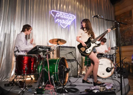 Band performing on a small stage with a metallic curtain backdrop and a neon 'prada mode' sign; keyboardist left, guitarist center, drummer right.