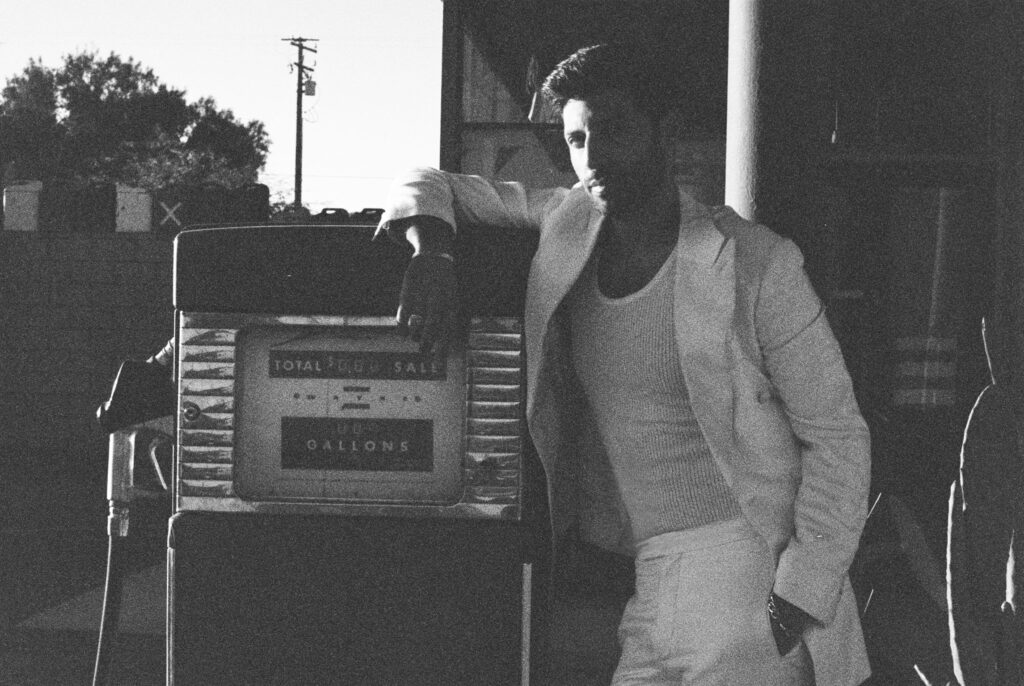 Joseph Reuben wearing a blazer and knit sweater leans on a vintage metal vending machine outdoors.
