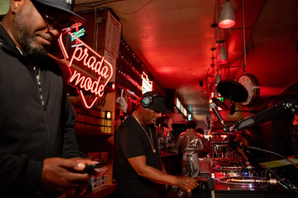 DJ in a black shirt and cap spinning records at a turntable in a neon red-lit bar scene, crowd in the background
