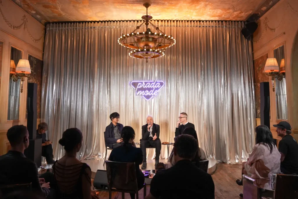 Panelists seated on a stage in front of silver-draped curtains, Prada Mode neon sign glowing above, audience in foreground.