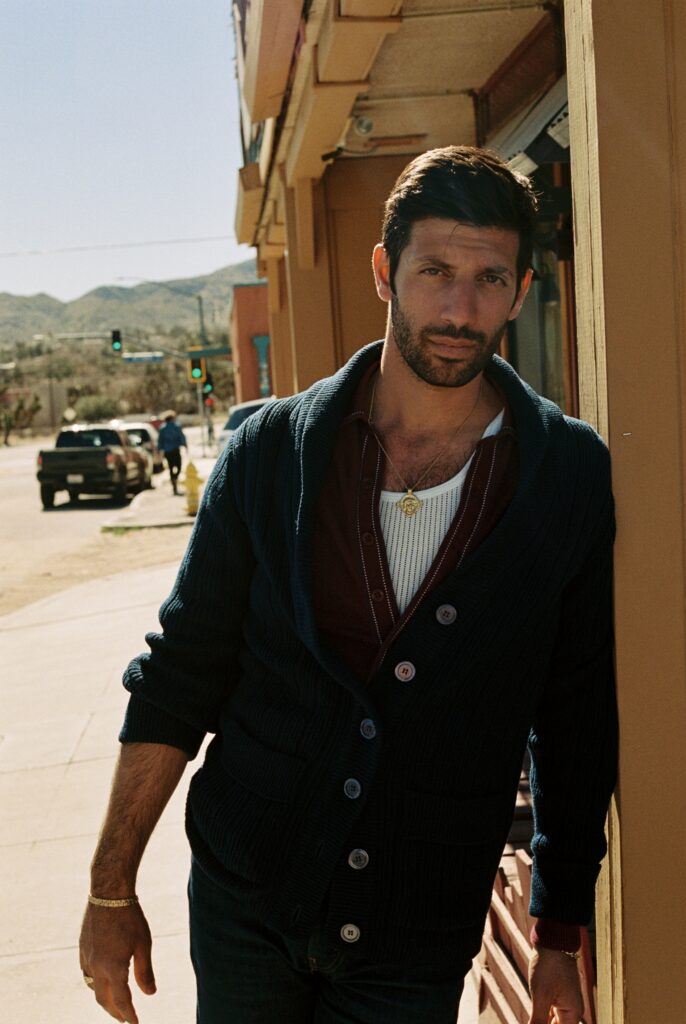 Joseph Reuben with dark hair and beard leaning against a beige building on a sunny city street, wearing a navy cardigan and gold necklace, with hills in the background.