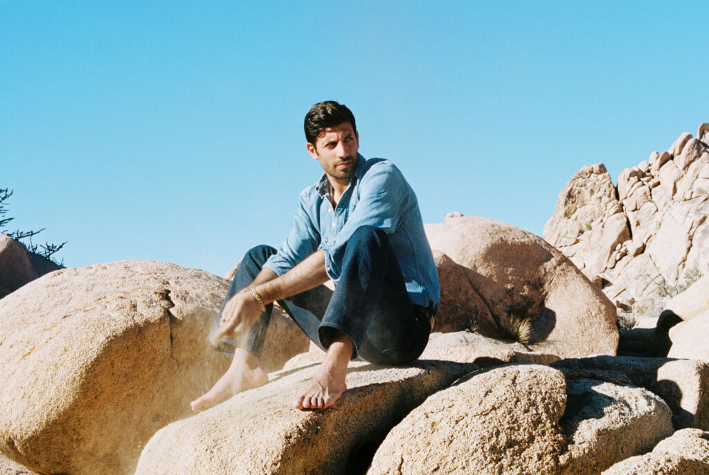 Joseph Reuben sitting on large desert rocks under a clear blue sky, wearing a light blue denim shirt and jeans, barefooted.