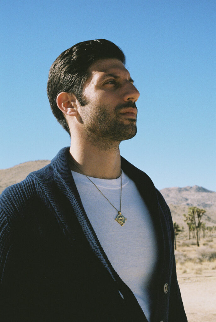 Portrait of Joseph Reuben outdoors in a blue sky setting, wearing a white shirt and dark cardigan with a pendant necklace, looking to the right.