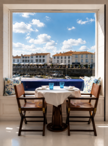 Table for two set with white tablecloth and blue glasses by a large window overlooking a seaside town and boats under a blue sky at le restaurant George's Île de Ré
