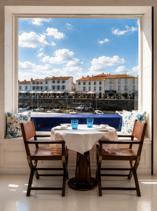 Table for two set with white tablecloth and blue glasses by a large window overlooking a seaside town and boats under a blue sky at le restaurant George's Île de Ré