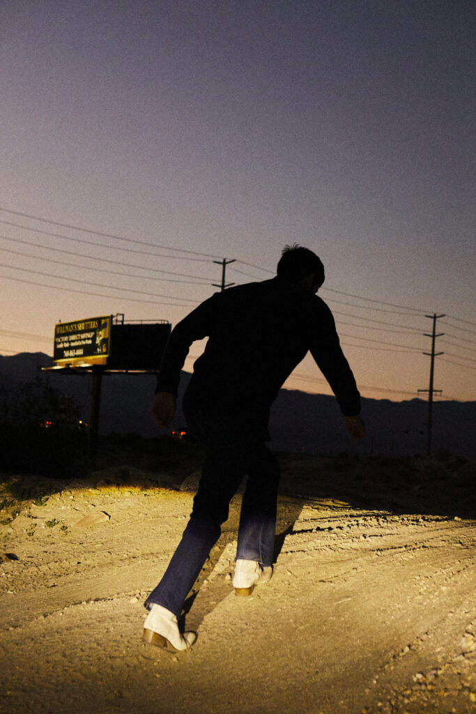 Silhouette of Joseph Reuben walking on rocky ground at dusk with a billboard and power lines in the background.