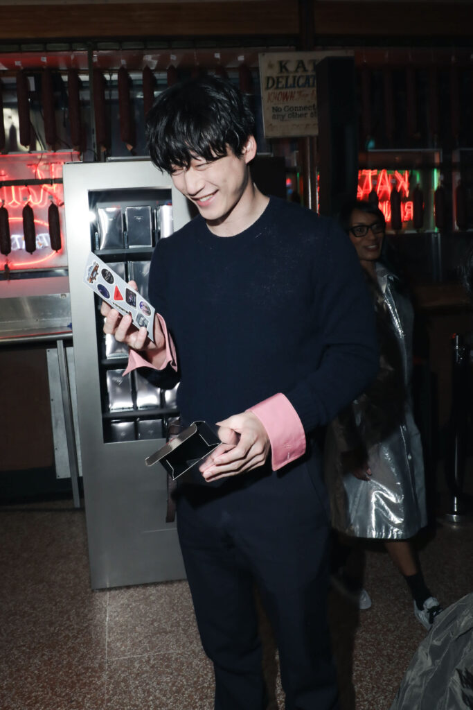 Young man in a navy sweater with pink cuffs smiles while inspecting a sheet of stickers in a neon-lit bar/arcade setting.