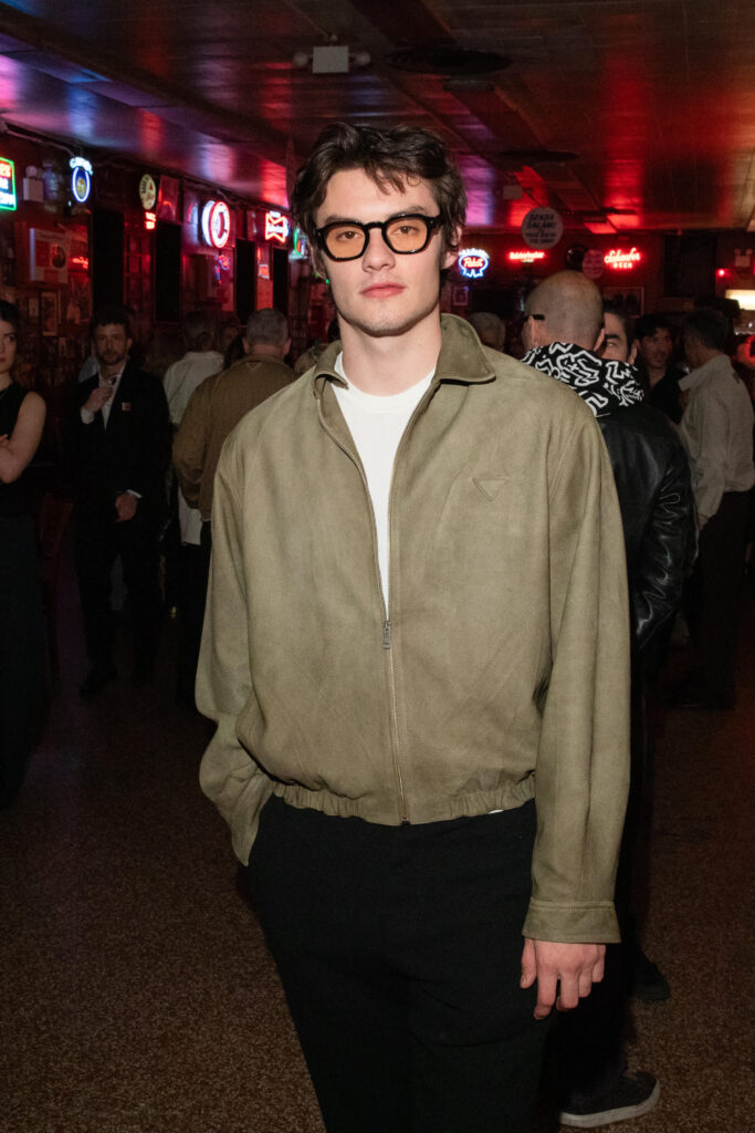 Portrait of a man with dark hair and black glasses, wearing a tan zip-up jacket, in a neon-lit bar crowd behind him.