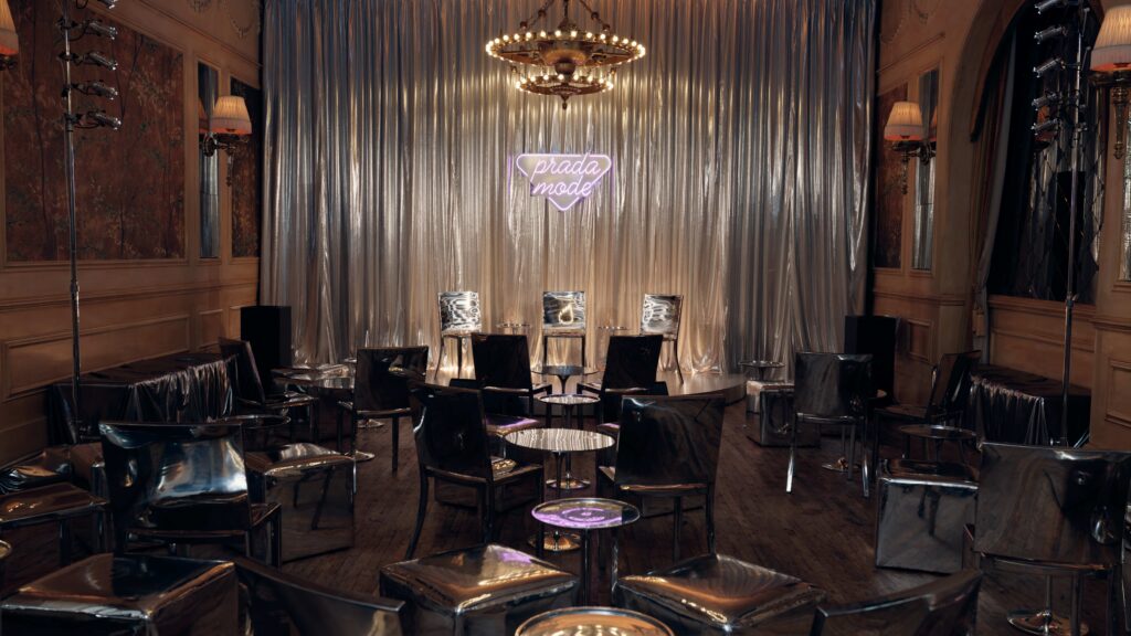 Stage setup with silver metallic chairs and round tables in a dim room, a silver curtain backdrop, and a pink neon Prada logo sign above the stage pieces.