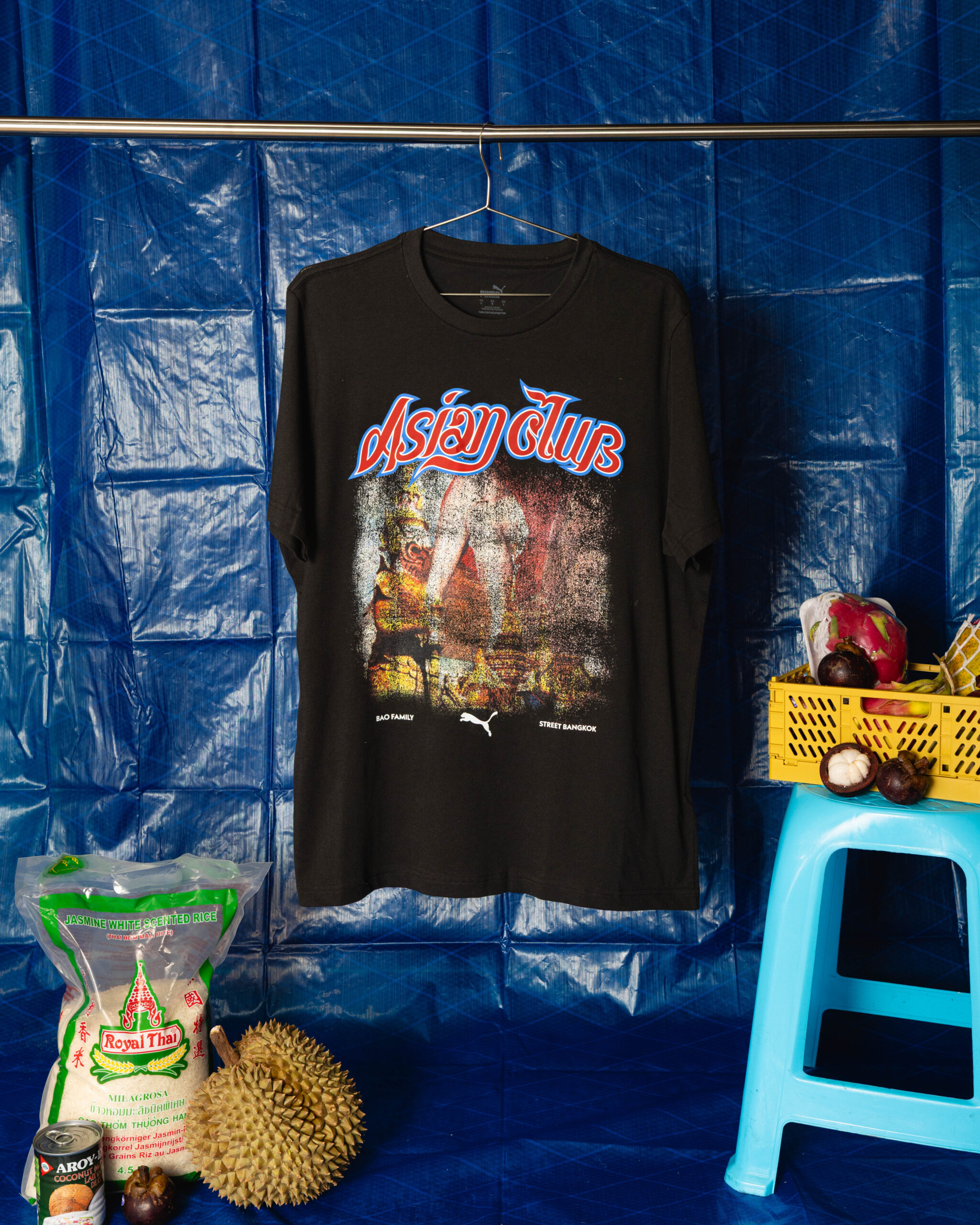 Black T-shirt with a red-and-blue 'Asia Club' graphic hanging on a wire hanger against a blue tarpaulin background; a yellow crate of fruit sits to the right.