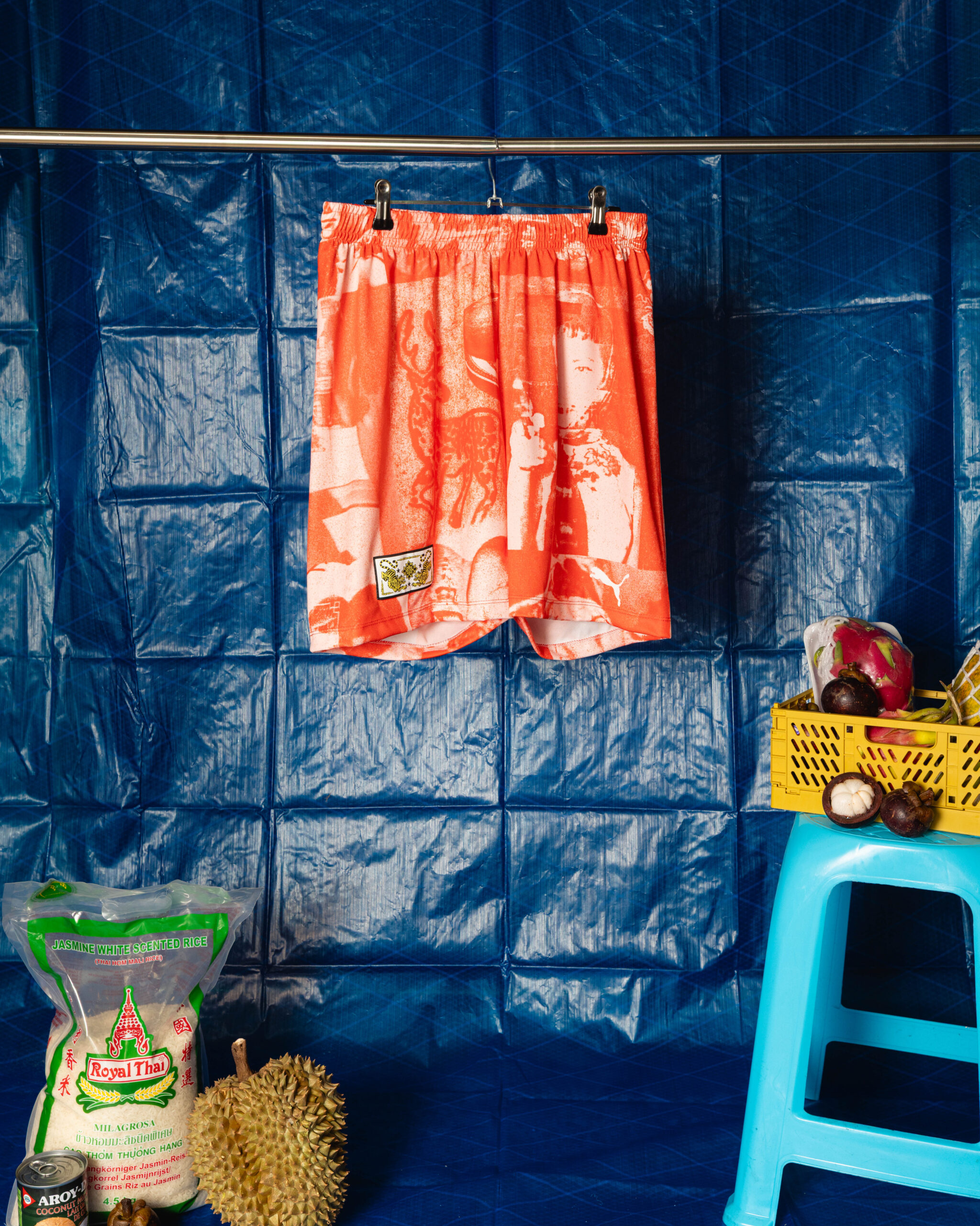 Orange patterned shorts hang on a clothesline against a blue tarp wall in a market setup, with a yellow crate of produce nearby and a bag of rice on the floor.