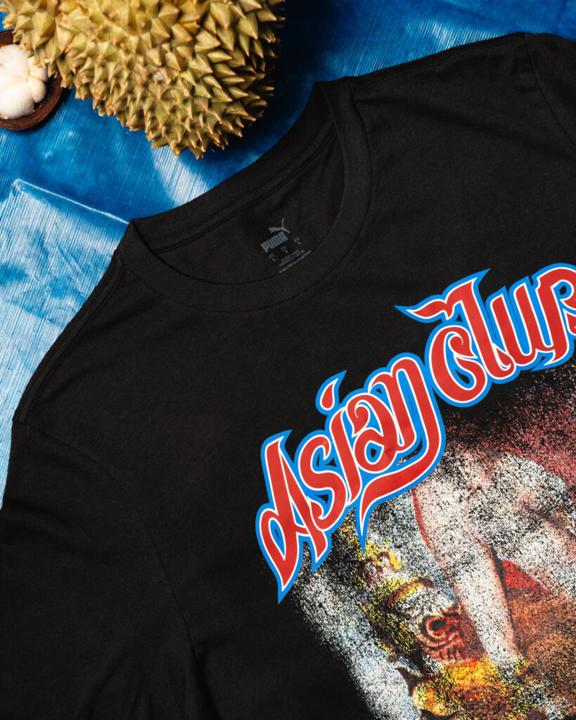 Close-up of a black T-shirt with a bold red-and-white graphic on the chest, set on a blue surface with a spiky durian fruit in the top left corner.