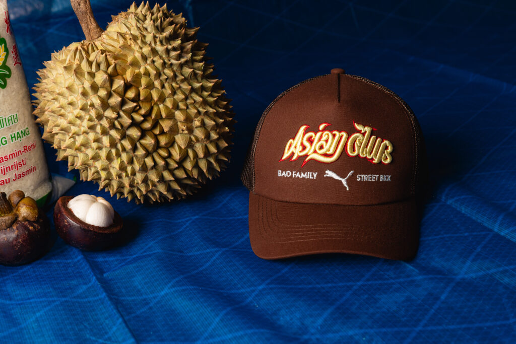 Spiky durian fruit beside a brown cap with embroidered lettering on a blue fabric background, with a bag of white rice partially visible on the left.
