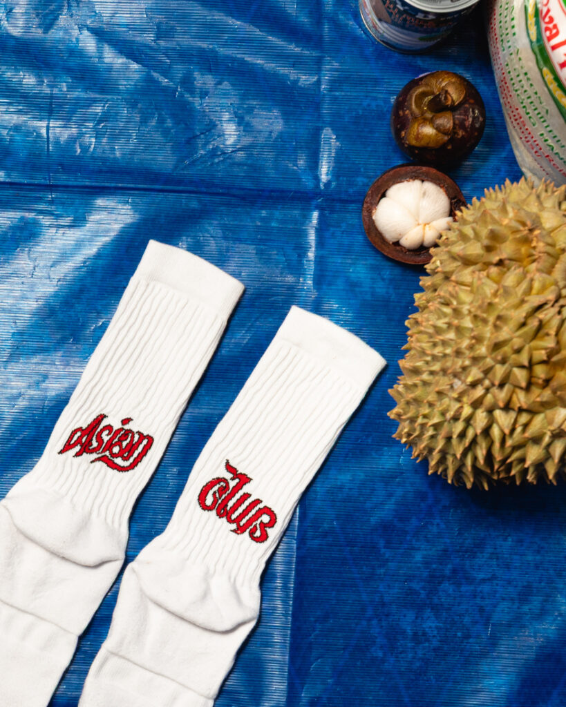 Pair of white socks with red embroidered text lying on a bright blue surface, near a durian fruit and its peeled white flesh in a shell.