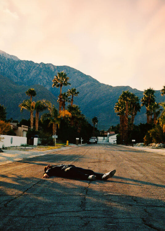Person lying face down on a quiet suburban street with palm trees and mountains in the background.