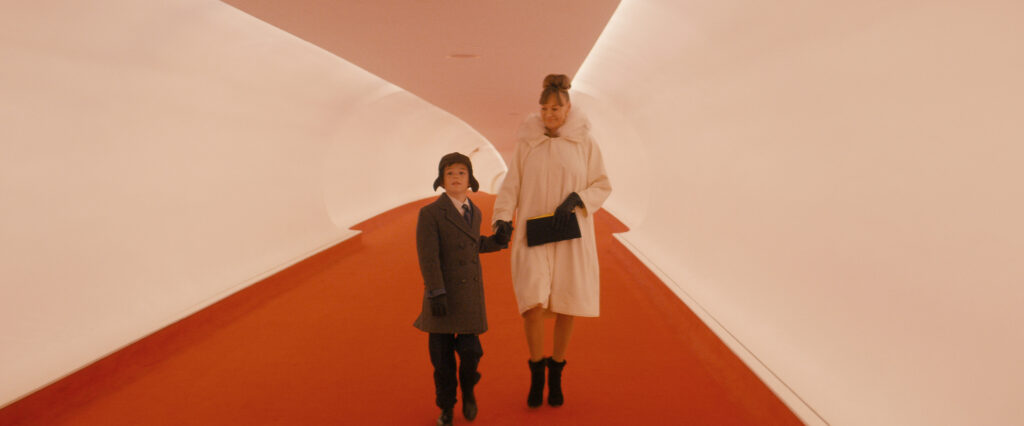 Woman in a white fur-collared coat and gloves holds a black clutch while walking hand-in-hand with a boy in a gray coat along a red carpeted hallway.