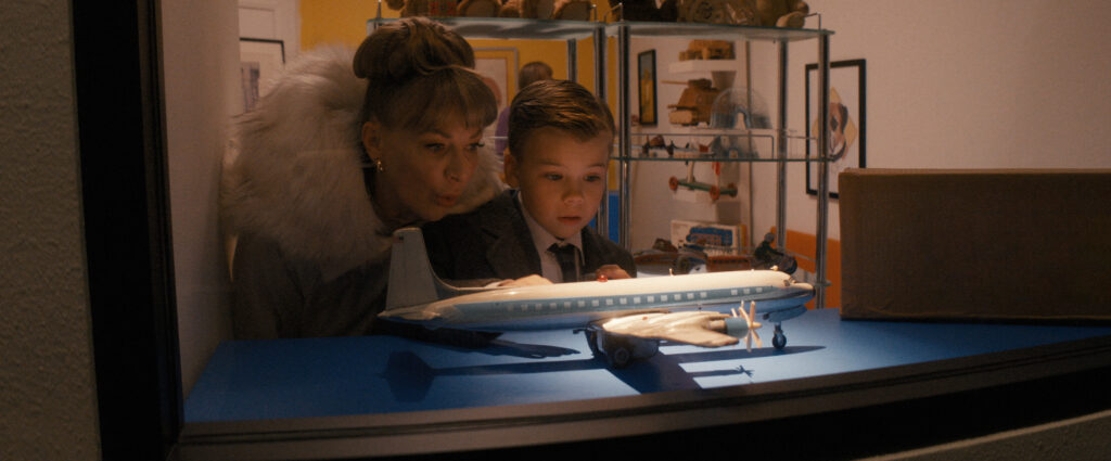 Woman with a fur stole leaning over a boy, both in formal clothing, examining a blue model airplane in a display case window.