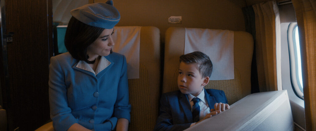 Flight attendant in a blue uniform smiling at a boy in a suit seated in an airplane cabin.