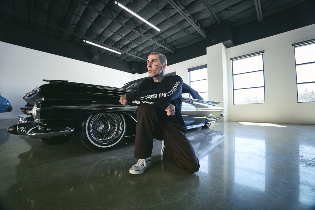 Man with tattoos kneels beside a glossy black vintage car inside a bright showroom or studio, looking off to the side.