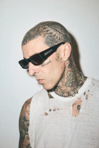 Side profile of Travis Barker wearing black sunglasses and a thick silver chain necklace, looking downward.
