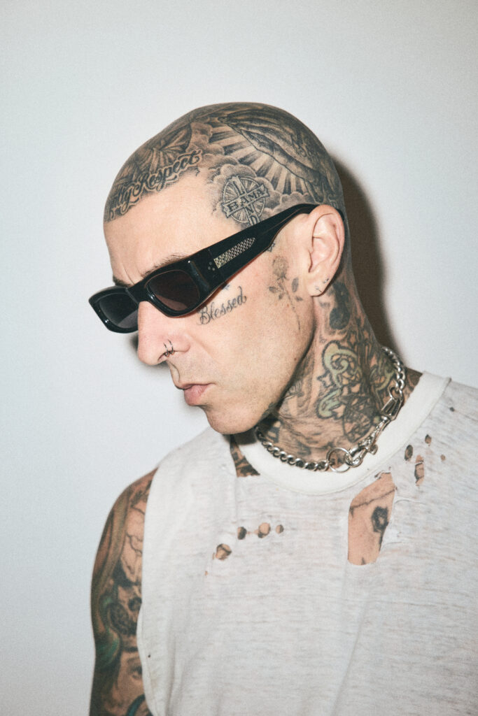Side profile of Travis Barker wearing black sunglasses and a thick silver chain necklace, looking downward.
