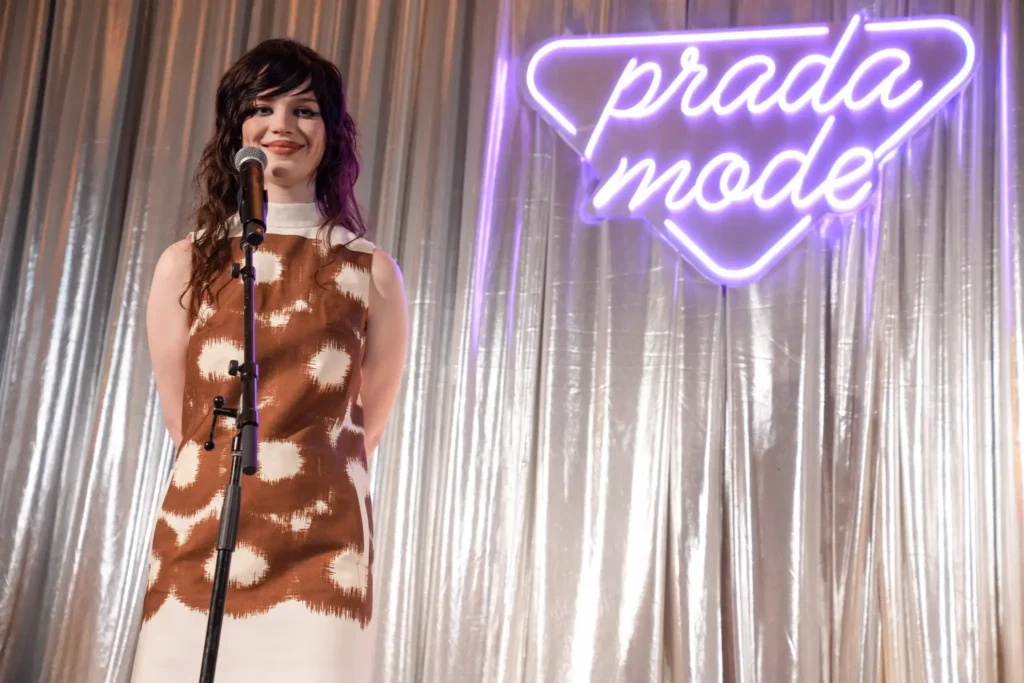 Woman with dark wavy hair stands at a microphone on a stage, wearing a brown and cream patterned dress, with a purple neon sign in the background.