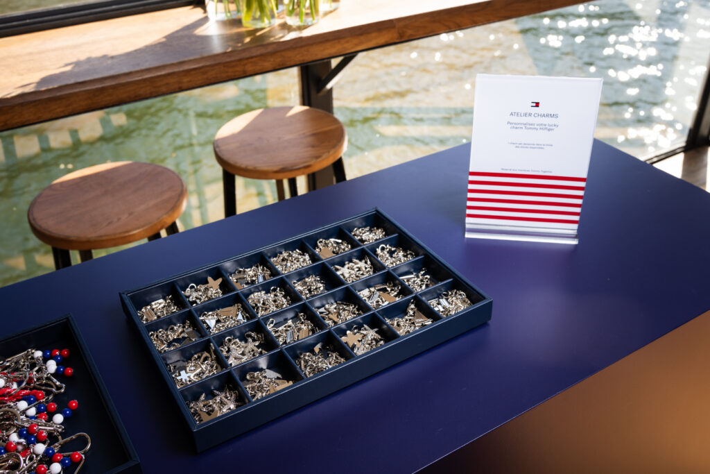 Display of metal charms and beaded keychain pieces in segmented blue tray on a purple table, with an Atelier Charms sign nearby by the water.