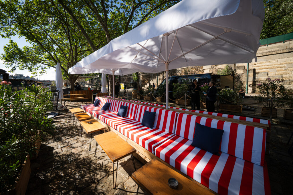 Outdoor dining area with white canopy tents, red-and-white striped cushioned benches and blue pillows along a stone-paved path
