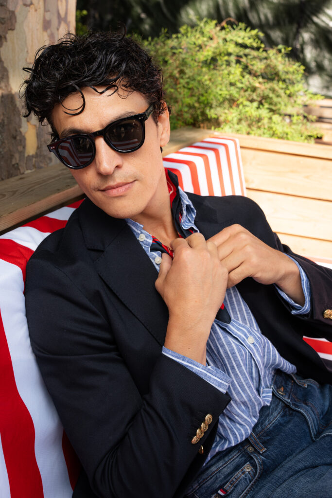 Man with dark sunglasses and curly hair lounges outdoors on a red-and-white striped cushion, wearing a navy blazer and striped shirt.