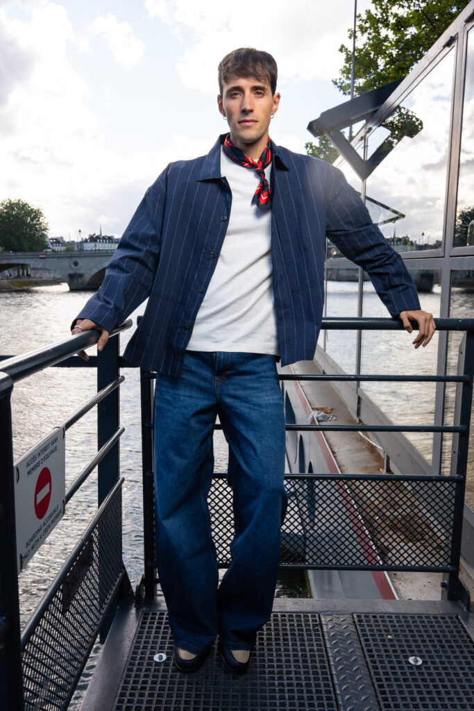 Man posing on a metal railing by a river, wearing a blue pinstripe jacket, white shirt, and red scarf, looking at the camera with a city bridge in the background.