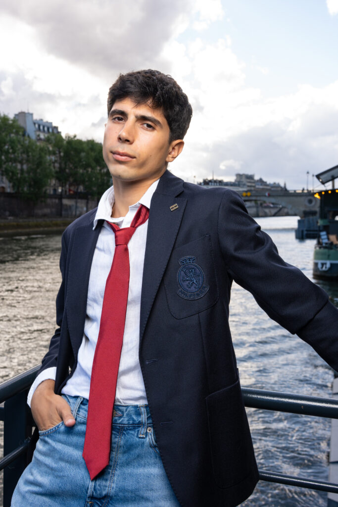 Young man in a navy blazer and red tie posing by a river with city buildings in the background