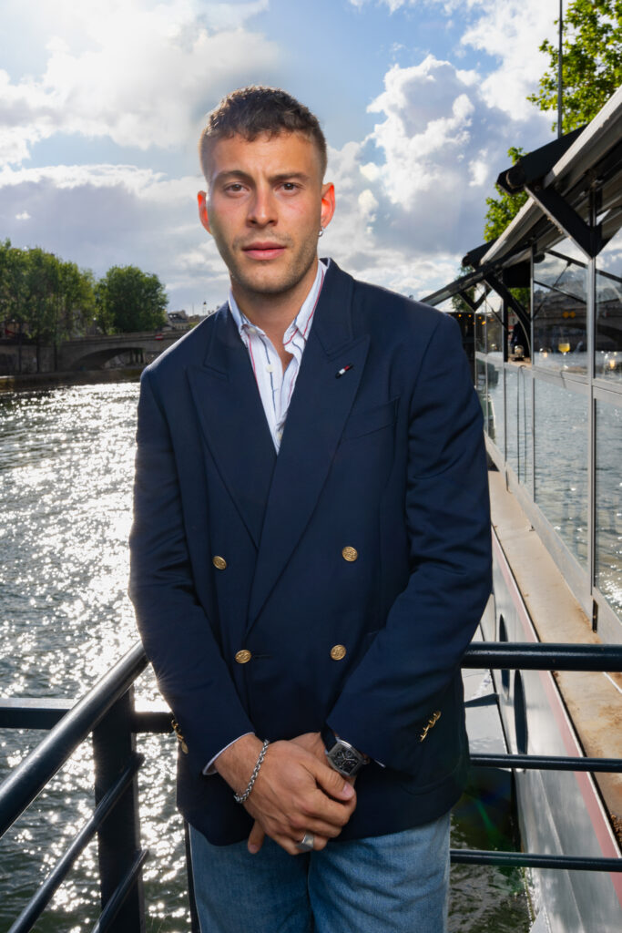 Ben Felipe in a navy blazer standing on a riverboat deck with water, trees, and a bridge in the background.