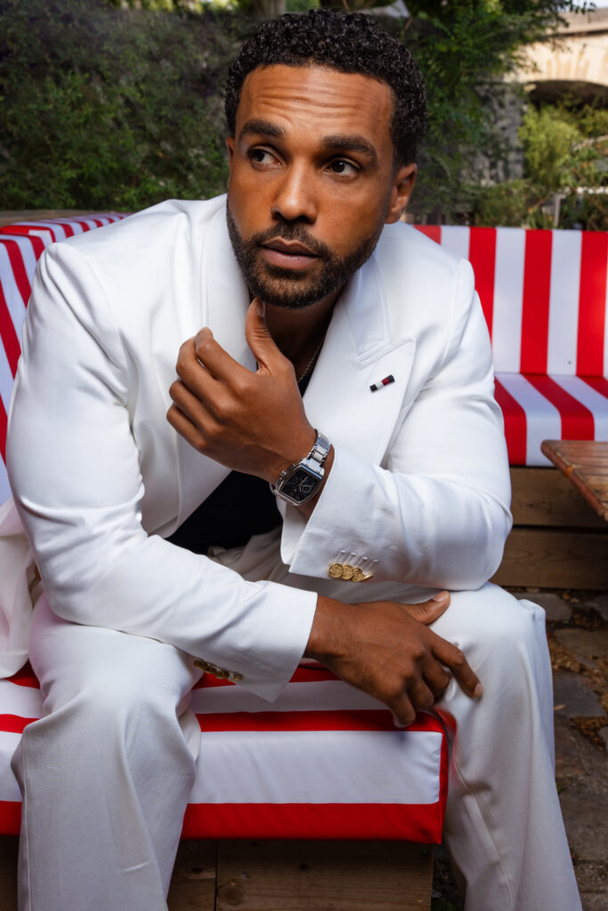 Lucien Laviscount in a white suit sits on a red-and-white striped bench outdoors, looking to the side, wearing a silver watch and gold cuff buttons.