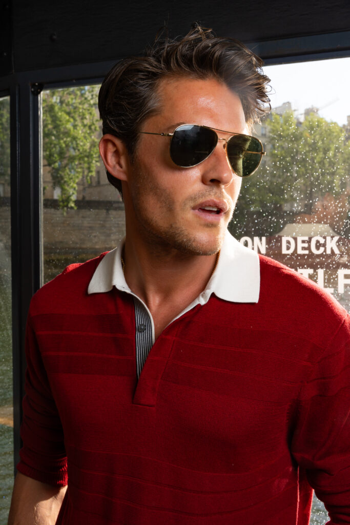 Man with dark hair wearing aviator sunglasses and a red polo shirt, looking off to the side by a window in daylight.