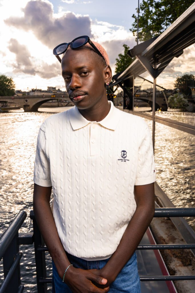 Portrait of a person with pink hair and sunglasses on their head, wearing a white polo sweater, standing by the river with a bridge in the background.