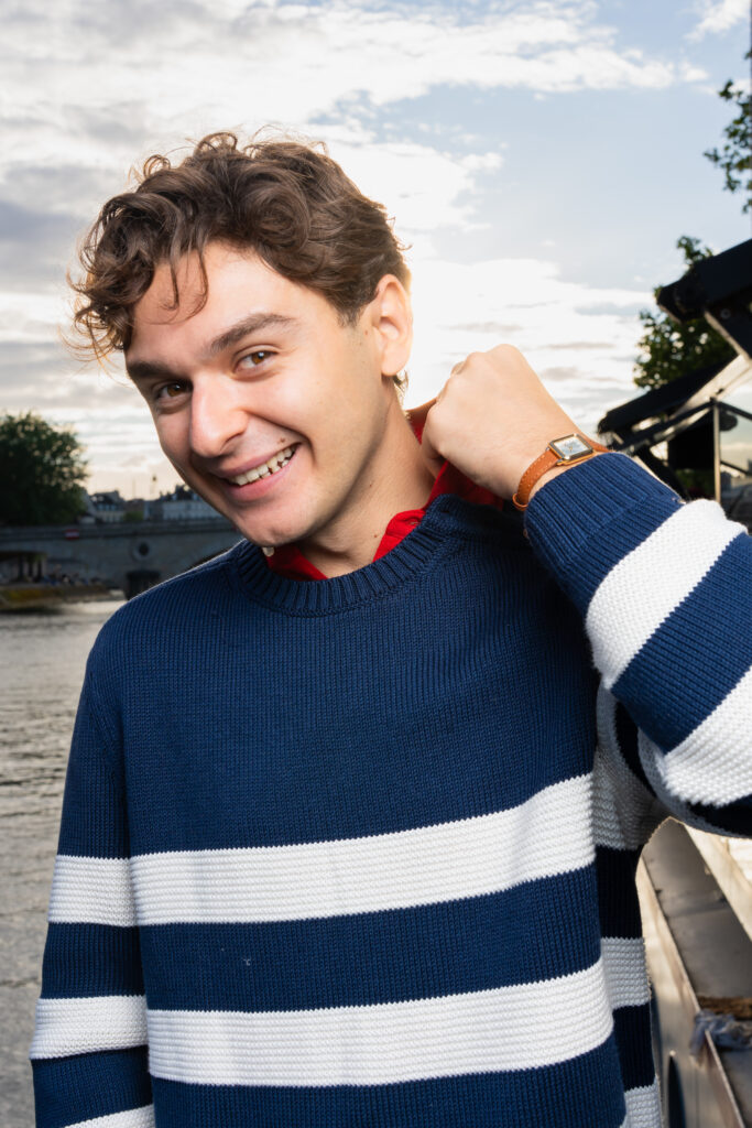 Young man smiling at camera outdoors by a river, wearing a blue and white striped sweater and a brown watch