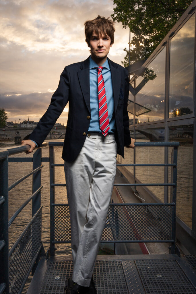 Young man in a navy blazer, blue shirt and red striped tie stands on a metal walkway by the river at sunset.