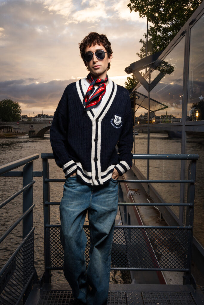 Young man in a navy cardigan with white stripes and red scarf stands on a railing by the river at sunset, wearing sunglasses.