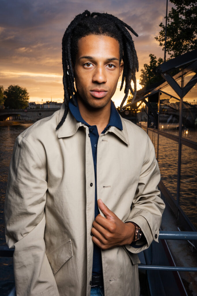Portrait of a young man with dreadlocks wearing a beige trench coat, standing on a riverside walkway at sunset.