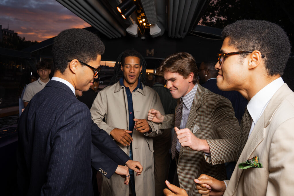 Group of young men in suits smiling and greeting each other at an evening outdoor party.